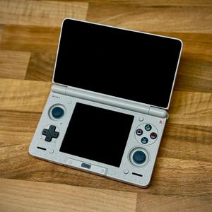 May include: A light gray handheld gaming console with two black screens, one open and one closed. The console has a directional pad, buttons, and two analog sticks. The console is on a wooden surface.