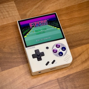 Può includere: Una console per videogiochi portatile bianca con un pad direzionale e pulsanti neri. Lo schermo mostra il gioco "F-ZERO" con il testo "Grand Prix Practice".