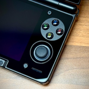 Pode incluir: Close-up de um console de jogos portátil preto. O console possui um direcional, um joystick e botões de ação coloridos. A tela é escura e o dispositivo tem um design elegante e moderno. O console está em uma superfície de madeira.