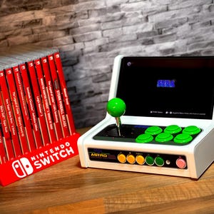 May include: A white and green retro arcade console with a joystick and buttons. The console has a black screen displaying the word "SEGA". The console is sitting on a wooden surface next to a red stand with the words "NINTENDO SWITCH" on it.