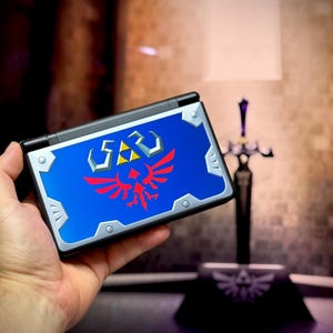 Puede incluir: Consola de juegos portátil con una cubierta azul con un emblema rojo y plateado. La consola se sostiene en una mano, con una espada decorativa en el fondo. El emblema incluye un escudo y una trifuerza.
