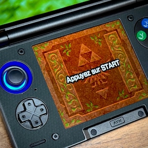 May include: Black handheld gaming console with a screen displaying a brown and green Zelda-themed graphic. The screen reads "Appuyez sur START". The console has blue-lit joysticks and coloured buttons.