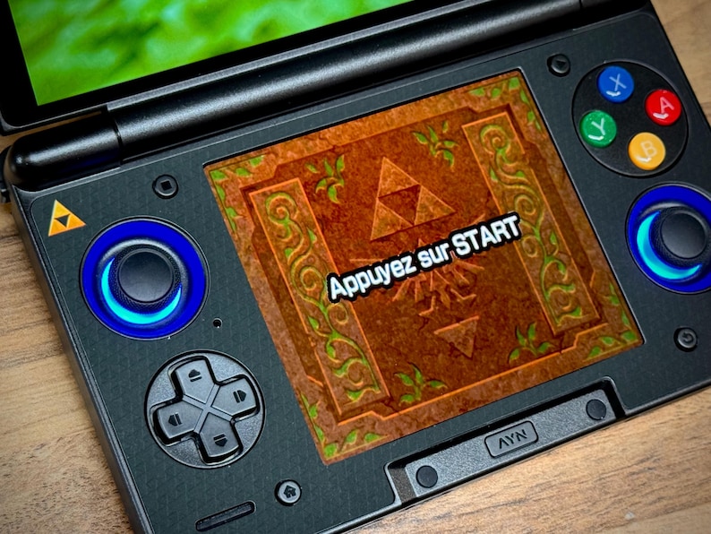 May include: Black handheld gaming console with a screen displaying a brown and green Zelda-themed graphic. The screen reads "Appuyez sur START". The console has blue-lit joysticks and coloured buttons.