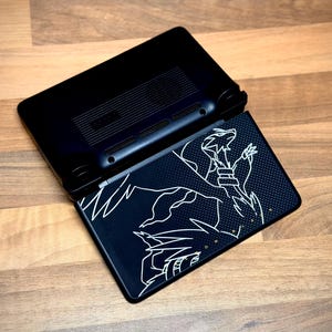 May include: Black handheld gaming console with a white graphic design on the bottom screen. The design features a dragon-like creature. The console is open, revealing the screen and buttons. The background is a wooden surface.