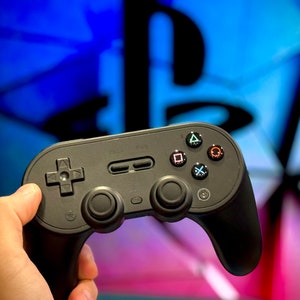 Peut inclure: Une manette de jeu sans fil noire avec un pavé directionnel, des boutons d'action et un bouton de menu central. La manette est tenue devant un fond bleu et blanc flou.