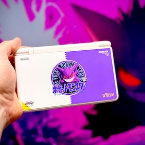 May include: A white and purple Nintendo DS Lite handheld game console featuring a Pokémon design. The design includes a purple Gengar character and Japanese text. The console is held in a hand against a purple and black background.