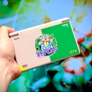 Peut inclure: Console de jeu portable Nintendo DS avec un design vert et beige. La console présente un autocollant avec un personnage Pokémon et du texte japonais. Le logo Game Boy est visible sur la console.