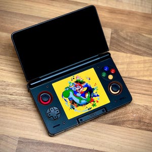 May include: Black handheld games console with a yellow screen showing a colourful cartoon image of Mario and Yoshi. The console has a black top screen and red and black buttons. The device is open on a wooden surface.