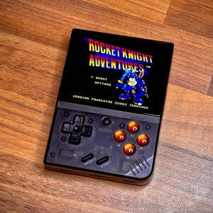May include: A black handheld video game console with a colour screen displaying the title "Rocket Knight Adventures" in a blue and red font. The console has a directional pad, four action buttons, and a select and start button.