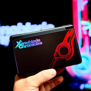May include: A black handheld gaming console featuring the Xenoblade Chronicles logo in a gradient blue and purple font on the left side, with a stylised red circular graphic design extending across the right side of the device's outer shell.