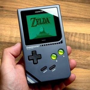 Op de afbeelding: Een grijze handheld gameconsole met een groen scherm met "The Legend of Zelda: Link's Awakening". De console heeft groene knoppen, een D-pad en de tekst "Chromatic" en "MOORETRO".