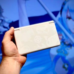 May include: A white handheld gaming console with a decorative pattern is held in a person's hand. The console is rectangular with a hinged top. The background is a blurred image of a blue-toned anime-style illustration.