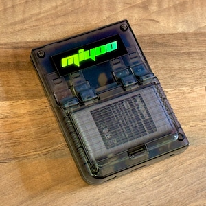May include: A black handheld gaming device with a transparent case. The device has a green and black logo that says "MiYOO".