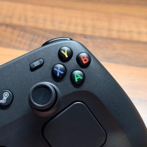 Op de afbeelding: Een zwarte videogamecontroller met een getextureerd oppervlak, een ronde joystick en vier hoofdactieknoppen met de letters Y, B, A en X in respectievelijk geel, rood, groen en blauw, naast een menuknop en een Steam-logo.