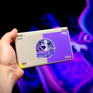 May include: A Nintendo Game Boy DS Lite console with a custom design featuring a purple and white graphic of a Pokemon character. The console has a gray and purple color scheme with Japanese text and a sticker.