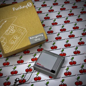 Puede incluir: Una consola port&aacute;til Funkey S Retrogaming gris con un logotipo de Nintendo Entertainment System negro y rojo. La consola est&aacute; sobre un fondo blanco con un patr&oacute;n repetitivo de cerezas rojas.