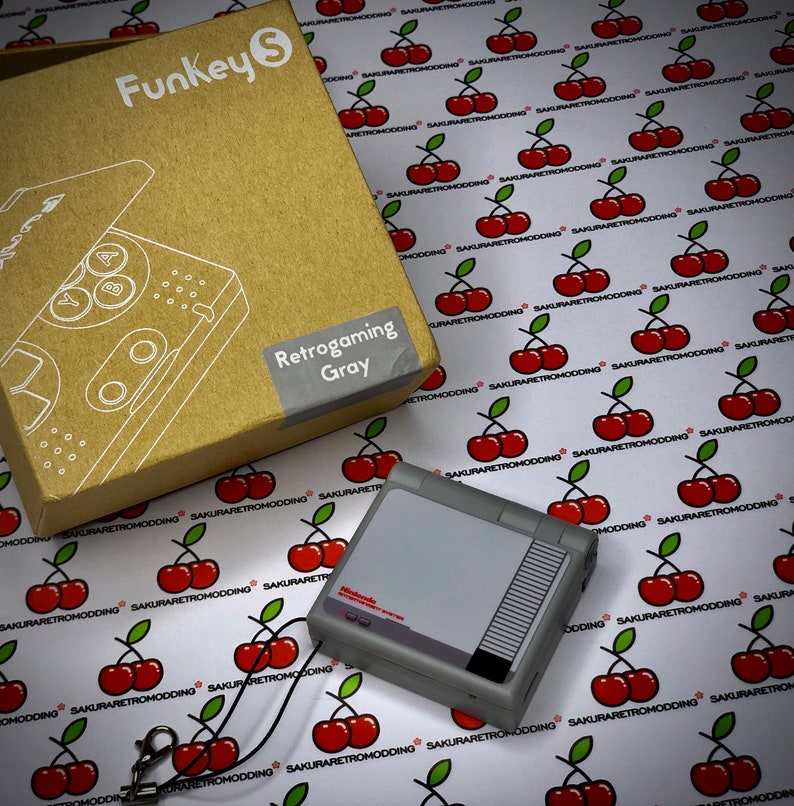 Puede incluir: Una consola port&aacute;til Funkey S Retrogaming gris con un logotipo de Nintendo Entertainment System negro y rojo. La consola est&aacute; sobre un fondo blanco con un patr&oacute;n repetitivo de cerezas rojas.