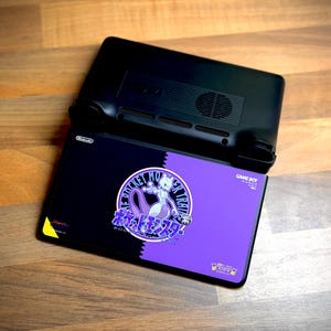 May include: Black and purple handheld gaming device with a custom design featuring a purple Mewtwo illustration and Japanese text. The device is open, revealing the screen with the design. The top portion is black.