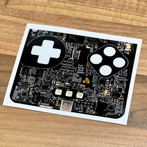 May include: A black and white circuit board with a white plus sign and four white circles. The board has text that reads "P/N: 6513C V0.06 060921".