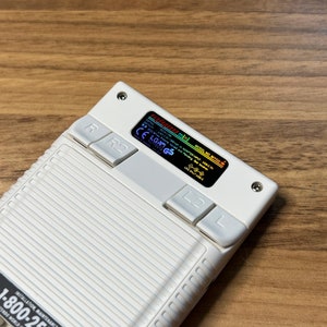 K&ouml;nnte beinhalten: Eine wei&szlig;e Handheld-Spielkonsole mit einem Schwarzwei&szlig;-Bildschirm, auf dem der Text "MODEL NO.  N64C-S" und "ADVANCED SPOT USB-C TV" angezeigt wird, sowie ein rot-blauer Knopf mit der Beschriftung "R" und "R2".