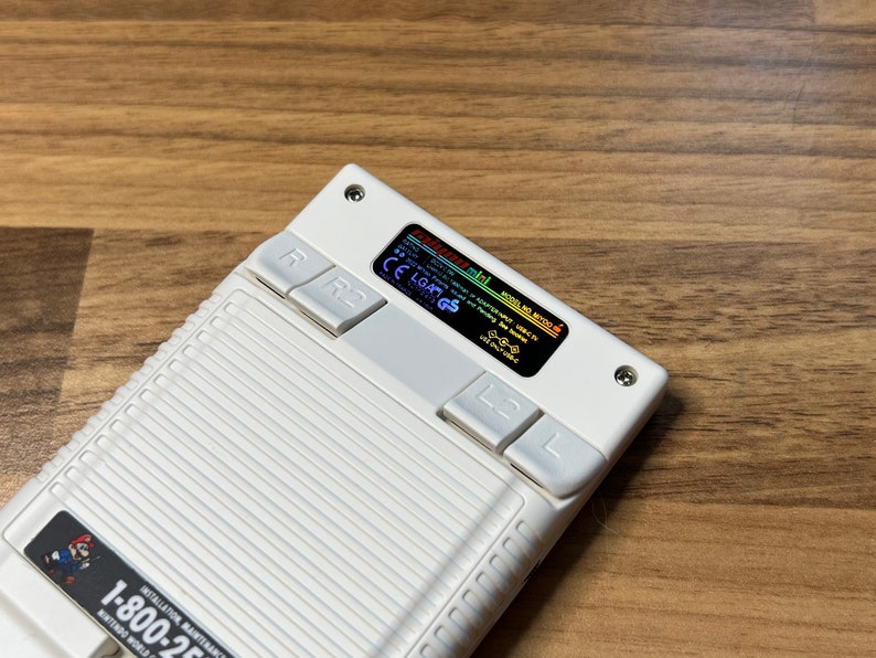 K&ouml;nnte beinhalten: Eine wei&szlig;e Handheld-Spielkonsole mit einem Schwarzwei&szlig;-Bildschirm, auf dem der Text "MODEL NO.  N64C-S" und "ADVANCED SPOT USB-C TV" angezeigt wird, sowie ein rot-blauer Knopf mit der Beschriftung "R" und "R2".