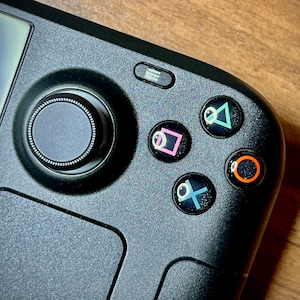 May include: Close-up of a black handheld gaming device with a circular control knob and four face buttons. The buttons are labelled with the symbols: triangle, square, circle, and cross.