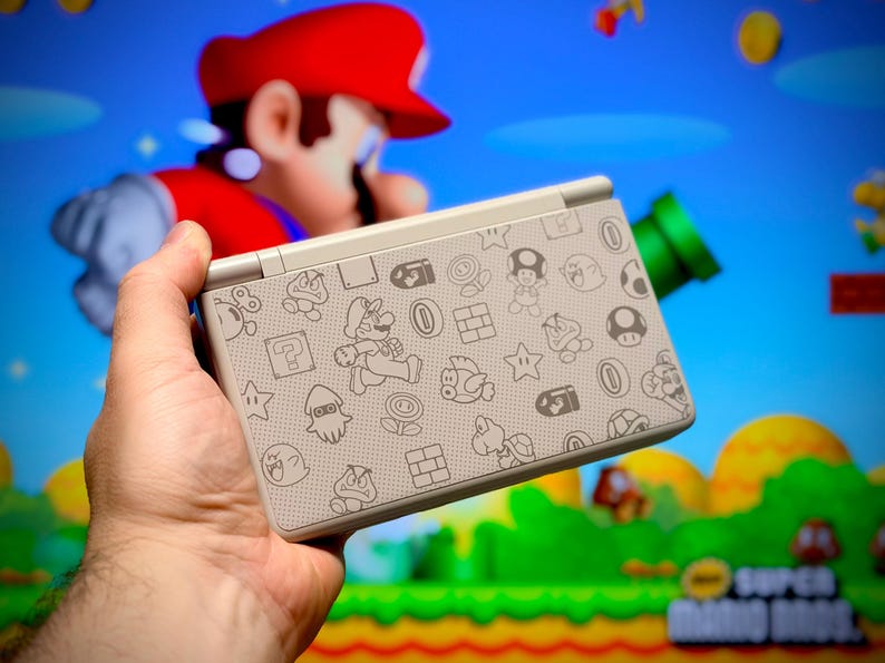 May include: A handheld video game console with a light gray cover featuring various cartoon character illustrations. The console is held in front of a blurred background of a Super Mario Bros. scene. The console's design includes icons of Mario, mushrooms, and stars.