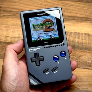 Op de afbeelding: Grijze handheld gameconsole met Super Mario Land 2 op het scherm. De console heeft een D-pad, blauwe knoppen en de tekst "CHROMATIC" en "MOORETRO". Het scherm toont een kleurrijk spel.