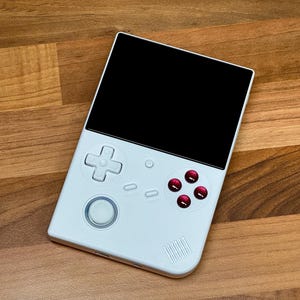 以下が含まれることがあります： 黒い画面、白い十字キー、白い丸いボタン、赤いボタンが4つ付いた白い携帯ゲーム機です。