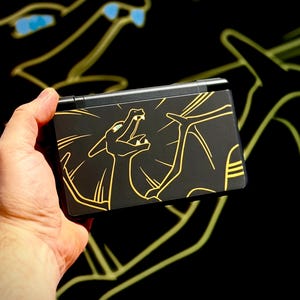 May include: Black handheld gaming console featuring a gold outline of a dragon-like creature. The console is held against a black background with a matching gold outline design. The console is closed and has a rectangular shape.