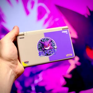 May include: A beige Nintendo DS Lite handheld game console with a purple and beige design. The console features a circular graphic of a purple Gengar character and Japanese text. The background is a blurred image of a purple Gengar.