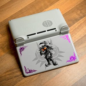 Op de afbeelding: Een lichtgrijze handheld gameconsole met een Majora's Mask-ontwerp op het onderste scherm. Het ontwerp heeft een karakterillustratie in zwart en grijs met oranje ogen en paarse decoratieve accenten. De bovenkant van de console is lichtgrijs met de tekst "NAVI".