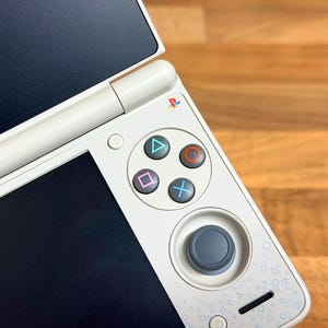 May include: A white handheld games console with a clamshell design. The console has a directional pad, action buttons, and a joystick. The screen is black. The console is on a wooden surface.
