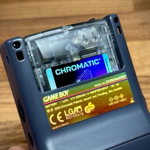Puede incluir: Primer plano de una consola Game Boy azul con una ranura transparente para cartuchos. El cartucho muestra la palabra "CHROMATIC" en blanco. La consola tiene una pegatina amarilla con texto que incluye "GAME BOY" y "MADE IN FRANCE".