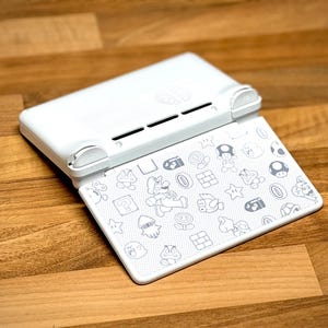 May include: White Nintendo Switch case, open to reveal a patterned interior. The interior features gray line drawings of characters and icons from the Super Mario video game series. The case is on a wooden surface.