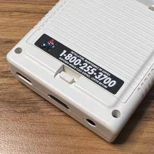 K&ouml;nnte beinhalten: Eine wei&szlig;e Handheld-Spielkonsole mit einem schwarzen Aufkleber, auf dem "Installation, Wartung oder Service 1-800-255-3700 Nintendo World Class Service Center Hotline" steht.