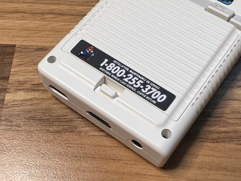 K&ouml;nnte beinhalten: Eine wei&szlig;e Handheld-Spielkonsole mit einem schwarzen Aufkleber, auf dem "Installation, Wartung oder Service 1-800-255-3700 Nintendo World Class Service Center Hotline" steht.