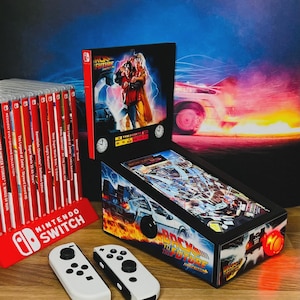 Op de afbeelding: Een Back to the Future-thema flipperkast met een bijpassende speldoos en twee witte Nintendo Switch controllers. De flipperkast heeft een kleurrijk ontwerp met het logo en de beelden van de film. Een rode Nintendo Switch spelhouder is zichtbaar.