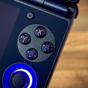 Puede incluir: Primer plano de una consola de juegos portátil. El dispositivo es azul oscuro con un panel de control circular y cuatro botones etiquetados X, Y, A y B en púrpura. Una luz azul rodea el joystick.