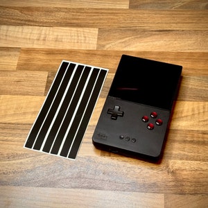 May include: A black handheld gaming console with a red button and a black and white screen. The console is on a wooden surface with a sheet of black stickers.