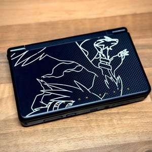 May include: Black handheld gaming console with a white graphic design of a dragon-like creature. The console is closed and resting on a wooden surface. The text "ON BLACK" is visible.