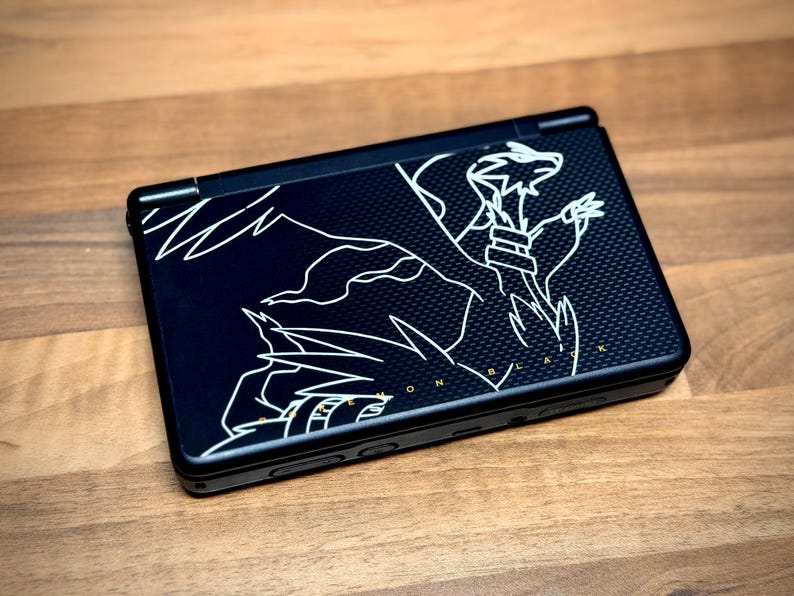May include: Black handheld gaming console with a white graphic design of a dragon-like creature. The console is closed and resting on a wooden surface. The text "ON BLACK" is visible.