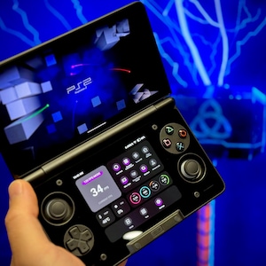 Puede incluir: Un dispositivo de juego portátil negro con una pantalla que muestra el logotipo "PS2" y un gráfico 3D. El dispositivo tiene múltiples botones, un joystick y una interfaz de pantalla táctil. La pantalla muestra una velocidad de fotogramas de 34 FPS.