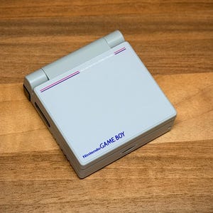 May include: A light grey Nintendo Game Boy Advance SP handheld console. The closed console displays the screen cover with the words "Nintendo GAME BOY" in blue. The top hinge is light grey with two thin purple and pink stripes.