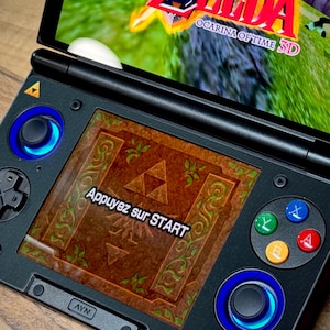 May include: A black handheld games console showing "The Legend of Zelda: Ocarina of Time 3D". The screen displays a game menu with the text "Appuyez sur START". The console has blue and coloured buttons.