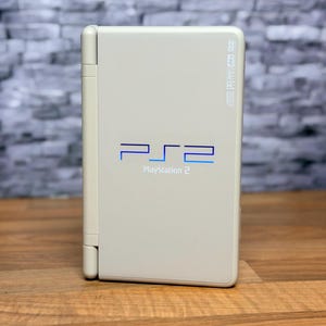 May include: A white PlayStation 2 console with the PS2 logo in blue. The console is standing upright on a wooden surface, with a gray brick wall in the background. The console has a DVD logo.