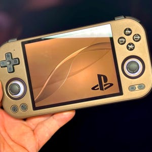以下が含まれることがあります： 金色の携帯ゲーム機が手に持たれています。画面には、曲線的なデザインとPlayStationのロゴが入った茶色の背景が表示されています。コンソールにはボタンとジョイスティックがあります。