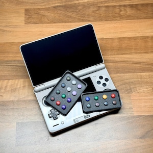 Peut inclure: Une console de jeu portable argentée avec un écran noir et une croix directionnelle, accompagnée de deux plateaux rectangulaires noirs contenant de petits boutons circulaires colorés dans des tons de rouge, bleu, vert, jaune et violet.