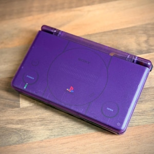 May include: A purple handheld gaming console with the Sony PlayStation logo and the words "POWER" and "OPEN" printed on the surface. The console is on a wooden surface.
