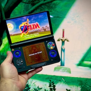 May include: A black Nintendo 3DS handheld game console showing a scene from "The Legend of Zelda". The top screen displays a castle and a logo. The bottom screen shows a game menu with the text "Appuyez sur START". A decorative sword is in the background.